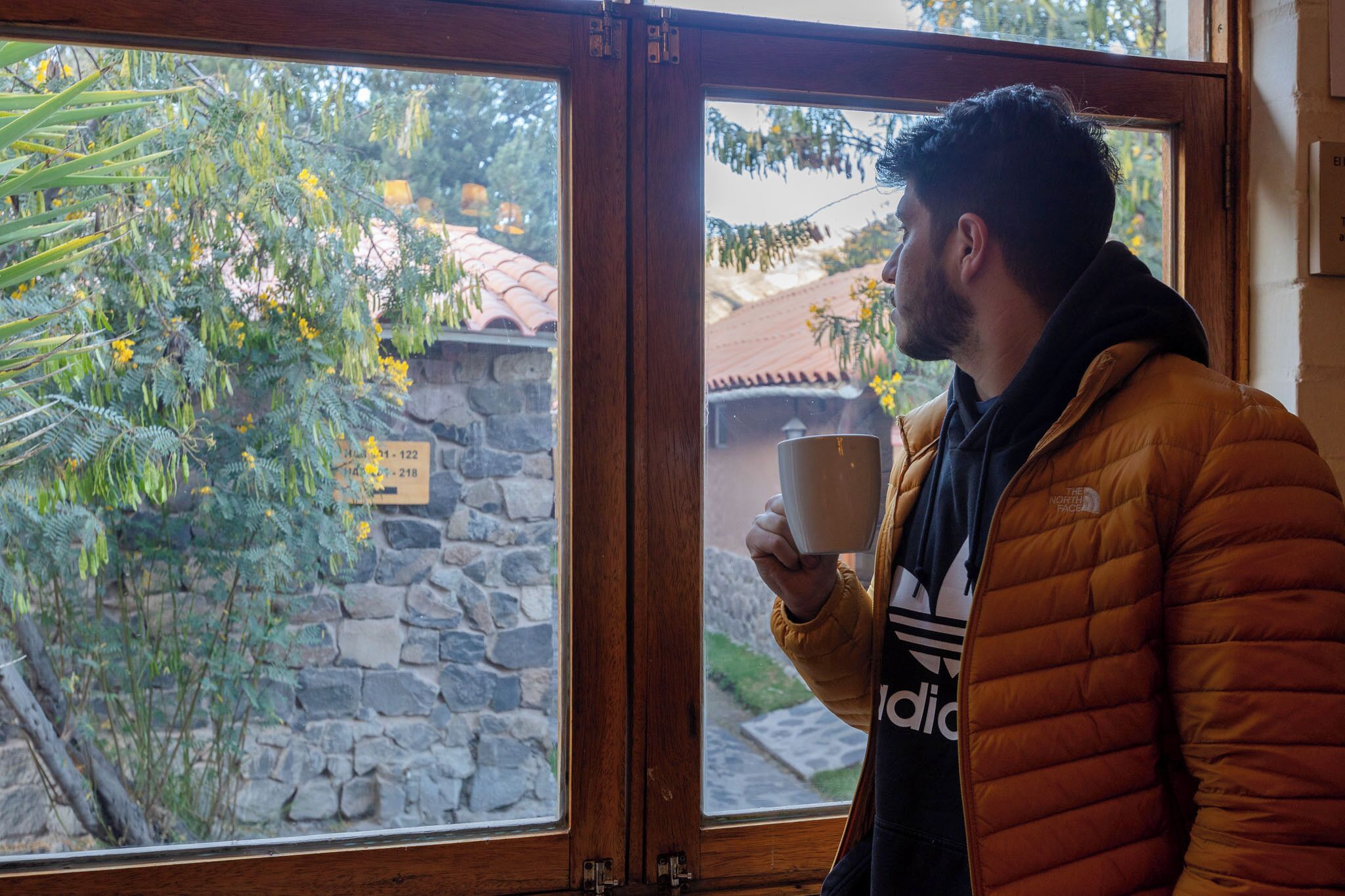 A person holding a cup near a window with a view of a stone wall and trees.