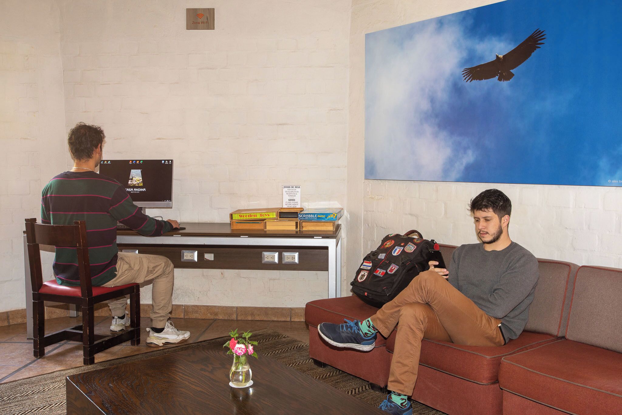 A man sitting on a couch and another man working at a desk with a computer. There is a painting of a bird on the wall, a vase with flowers on the table, and a backpack on the couch.