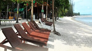 A beachside area with wooden lounge chairs and tables under trees, a building with a green roof, and a sandy beach. - Grand Sea View Resotel (Koh Samui)