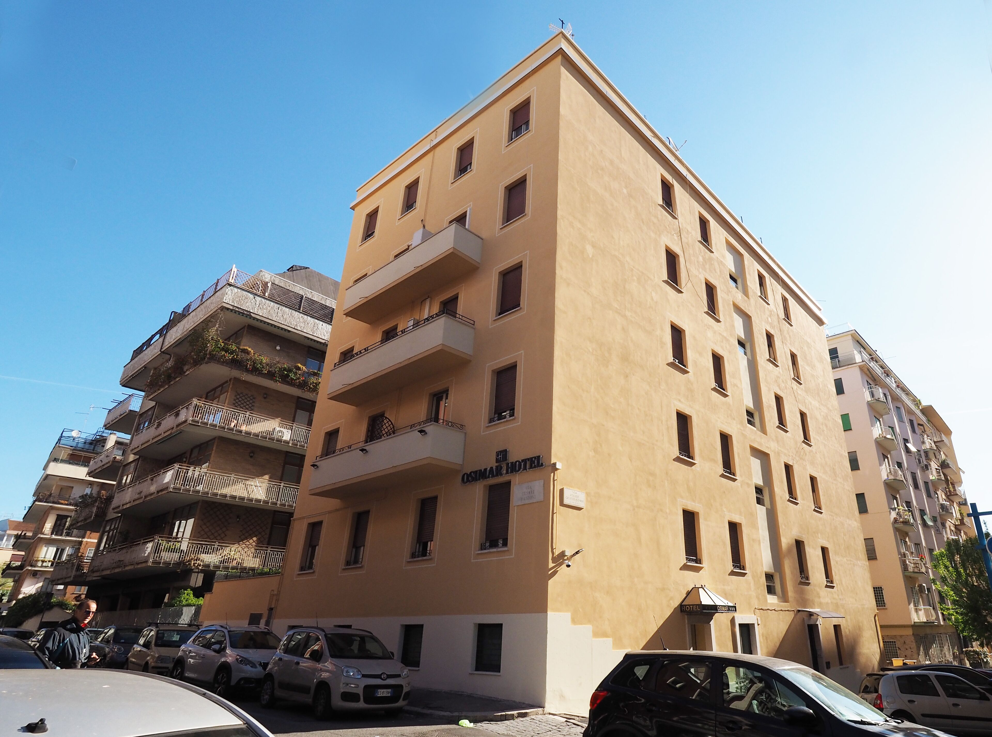 A multi-story hotel with balconies, a sign indicating it is the "OSIMAR HOTEL," and several parked cars in front.