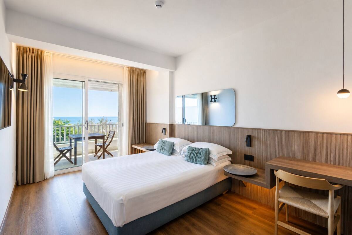 A hotel room with a large bed, a desk, a chair, a mirror, and a balcony with a view.