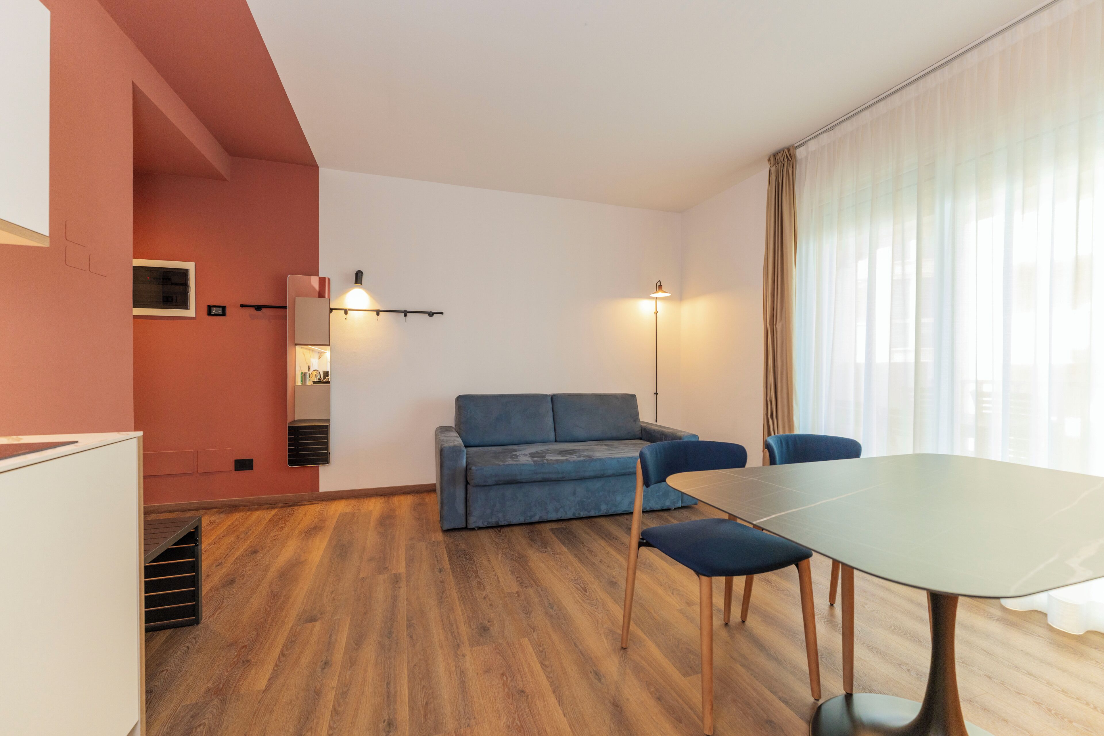 Ein Zimmer mit Holzboden, einem blauen Sofa, einem Tisch mit Stühlen, einer Stehlampe und einem fenster mit Vorhängen.