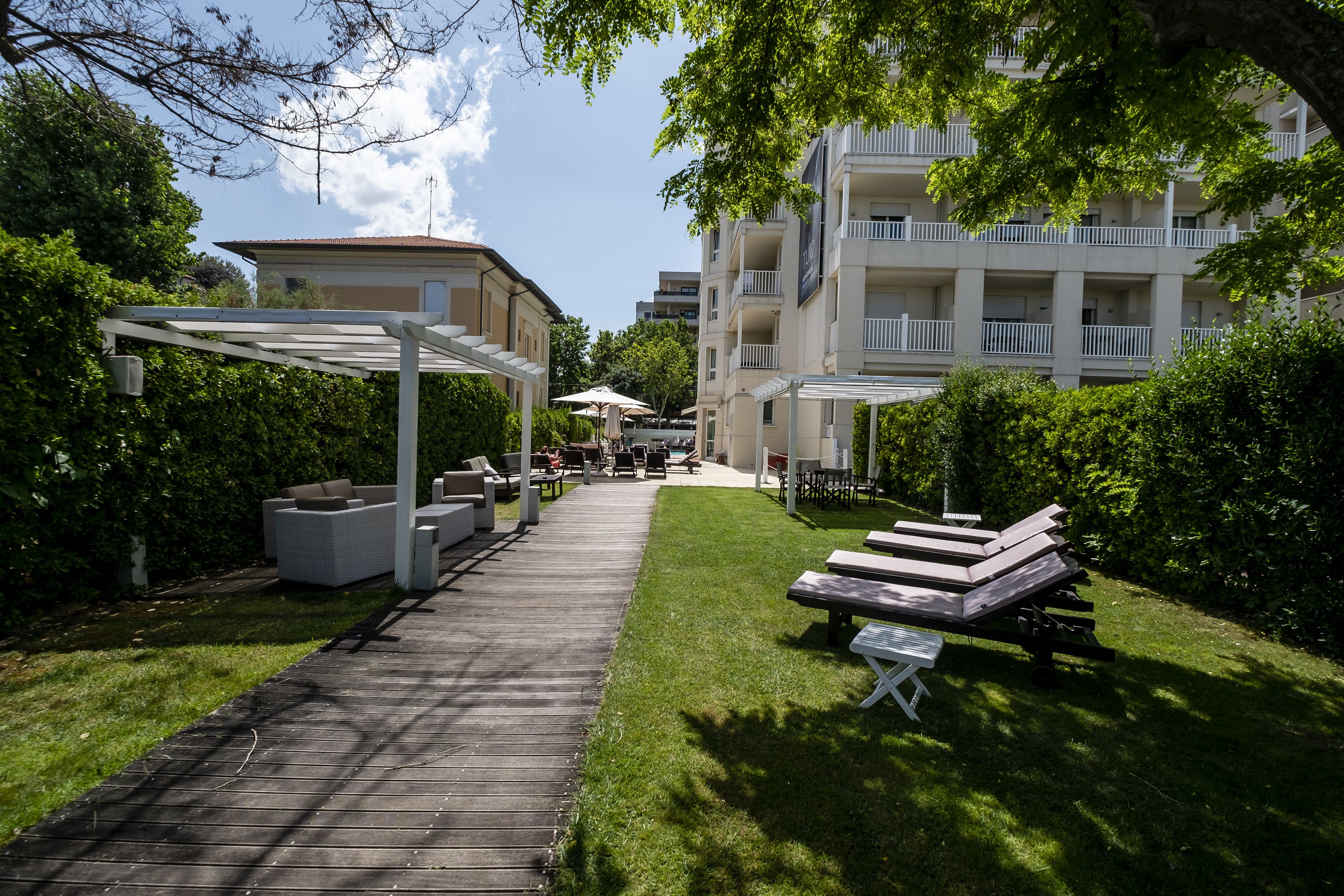 Una terraza de madera que conduce a un área de estar al aire libre cubierta, con sillones y sombrillas, rodeada de vegetación y un edificio de varios pisos con balcones.