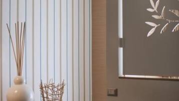 A room with vertical blinds, a vase with reeds, and a wall-mounted artwork.