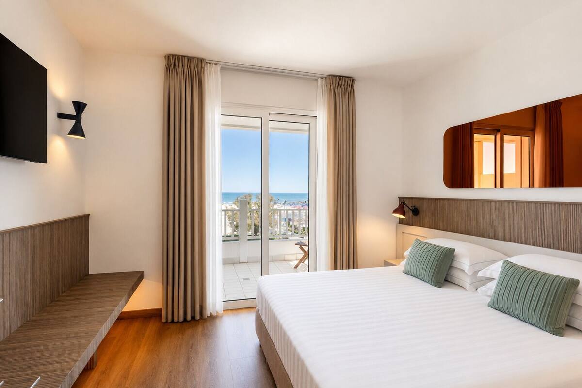 A hotel room with a bed, a TV, a balcony with curtains, and a view of the beach.