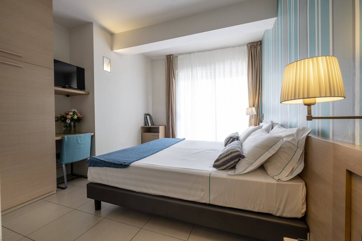 A modern hotel room with a bed, bedside table, lamp, TV, and a desk with a chair.