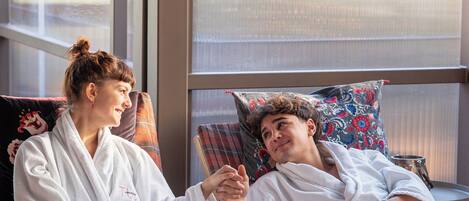 Two individuals in white bathrobes sitting on a wooden bench with a plaid cushion, holding blue mugs, in a room with large windows and a view of greenery outside.