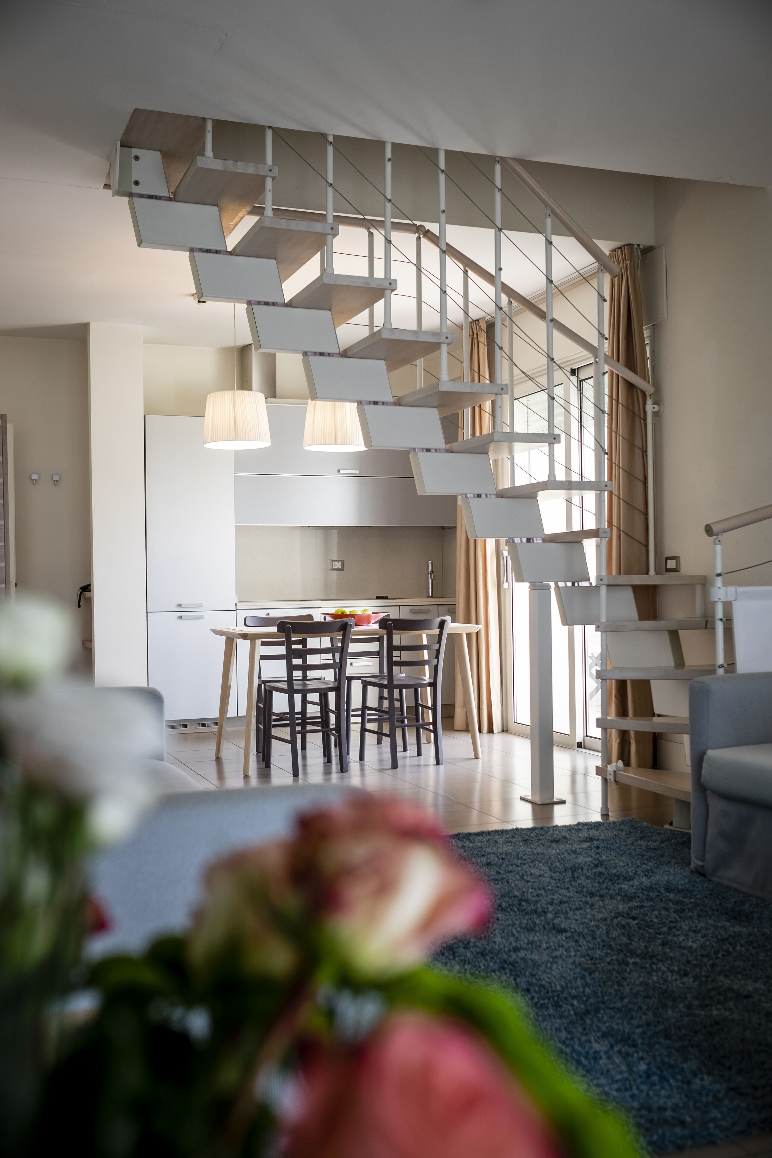 Un interno moderno con una scala che conduce a un piano superiore, un'area pranzo con tavolo e sedie, e un soggiorno con divano e tappeto blu.