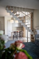 Un interno moderno con una scala che conduce a un piano superiore, un'area pranzo con tavolo e sedie, e un soggiorno con divano e tappeto blu.