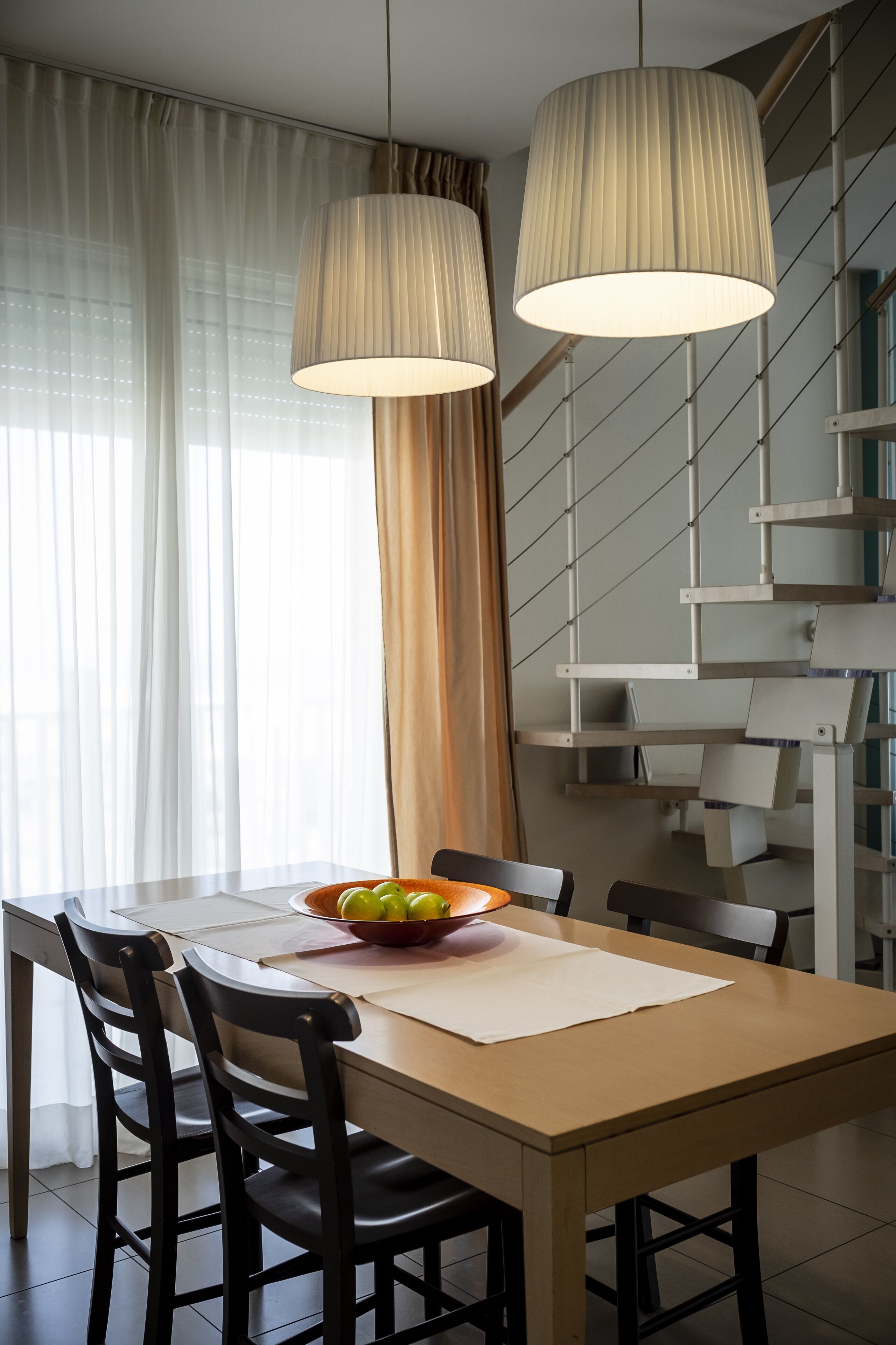 A dining area with a wooden table, black chairs, a bowl of fruit, and a staircase in the background.