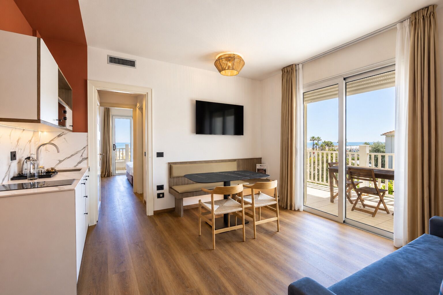 A modern hotel room with a kitchen, a dining area with a table and chairs, a sofa, a TV mounted on the wall, and a balcony with a view.