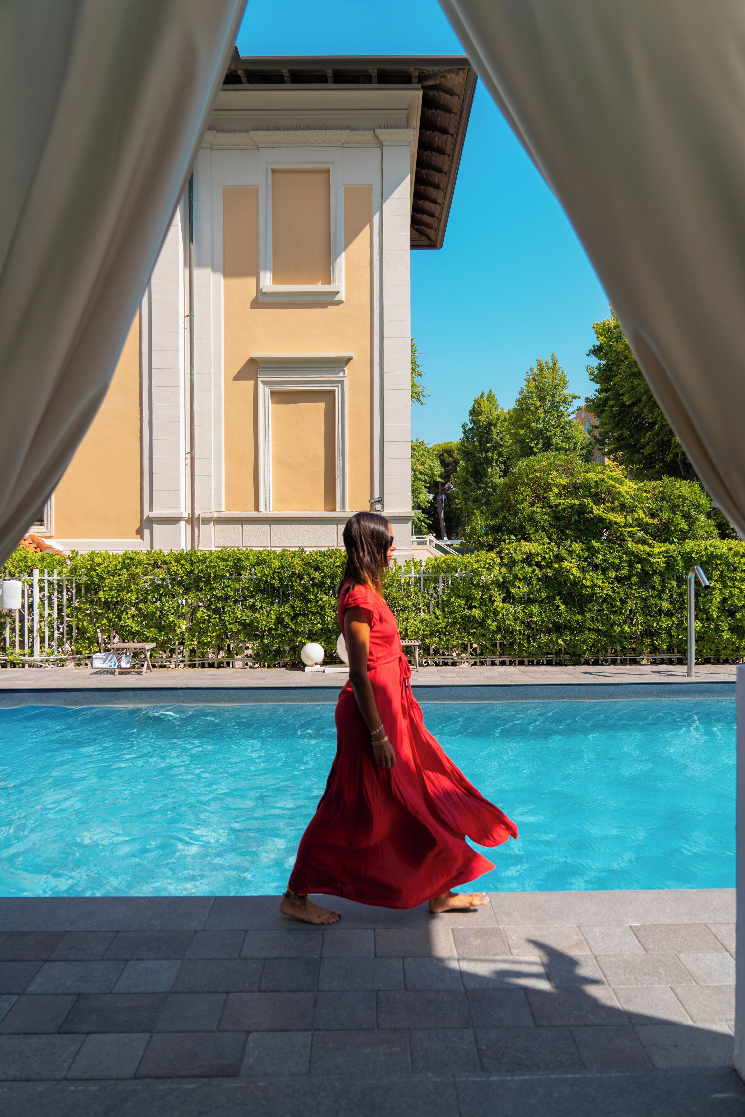 Mulher de vestido vermelho caminhando perto de uma piscina, com um prédio ao fundo.