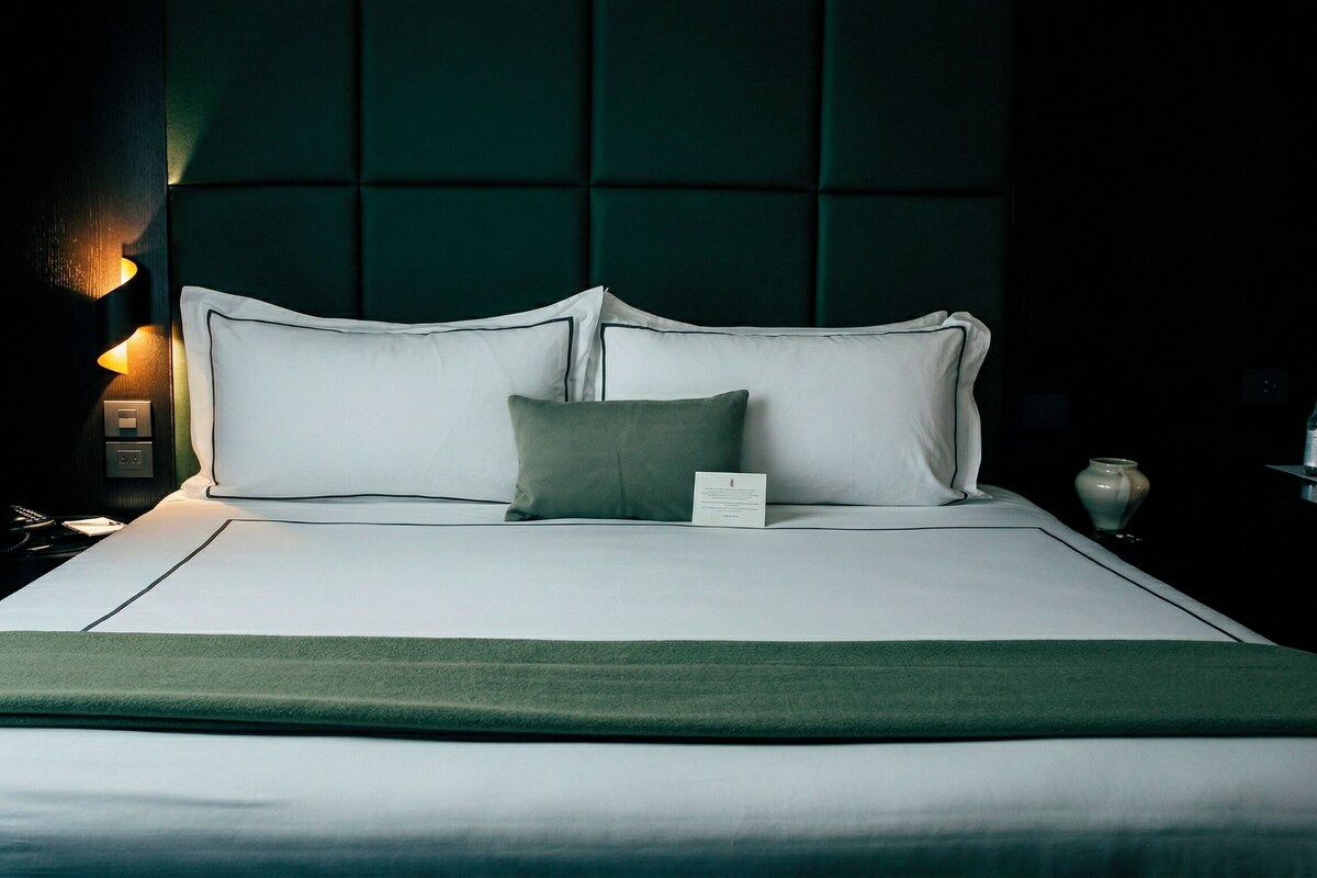 A neatly made bed with white linens and a green throw blanket, a bedside table with a lamp, and a dark green headboard.
