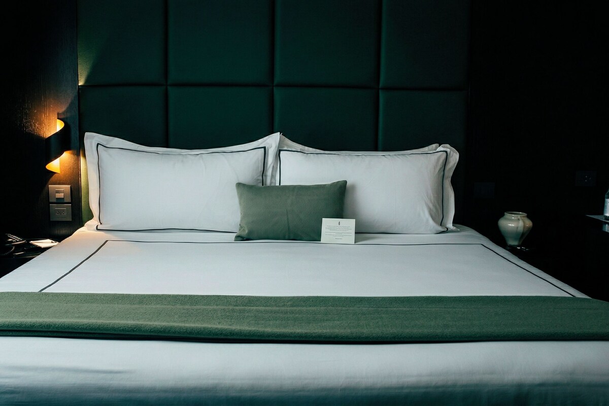 Una cama bien tendida con sábanas blancas y un plaid verde, una mesita de noche con una lámpara y un cabecero verde oscuro.