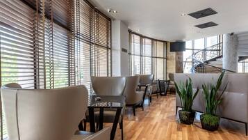 A spacious lounge area with wooden flooring, modern furniture, and large windows with blinds.