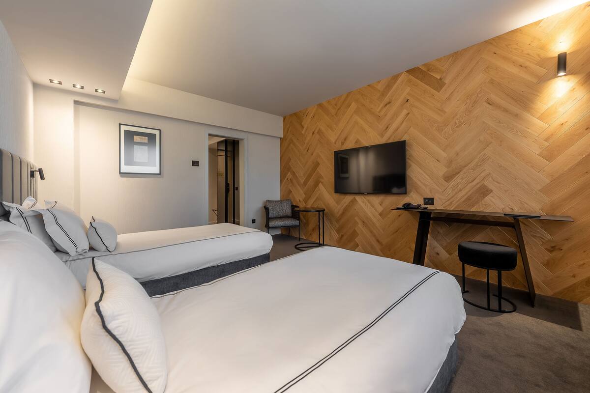 A modern hotel room with two beds, a desk, a chair, and a TV mounted on a wooden wall.