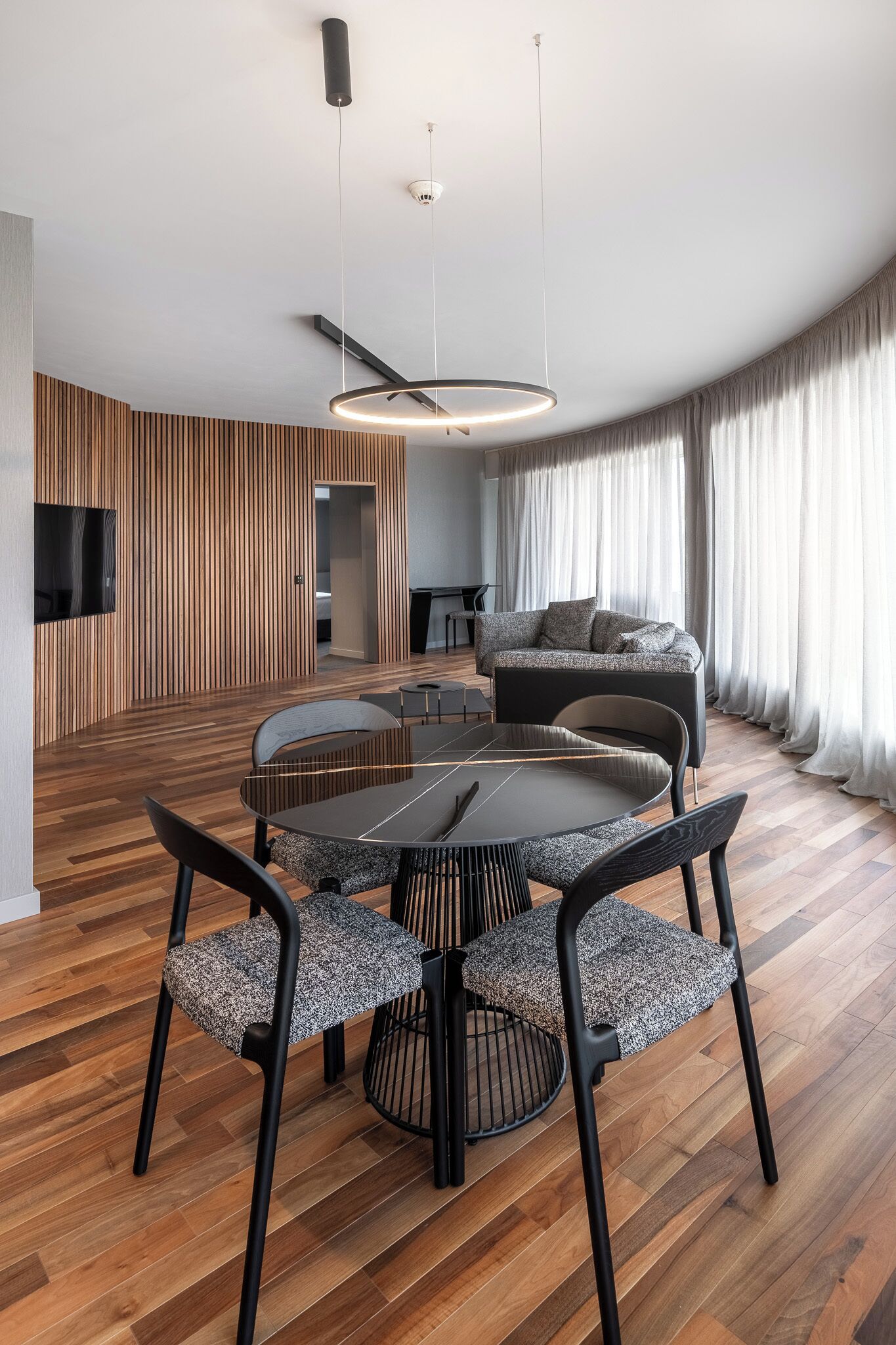 Een moderne woonkamer met een ronde eettafel, zwarte stoelen, een bank en een aan de muur bevestigde televisie.