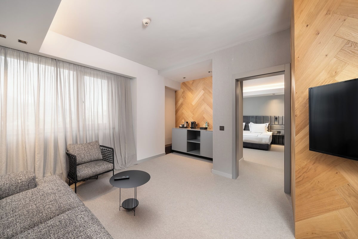 A modern hotel room with a sofa, a small table, a TV, a kitchenette, and a bedroom with a bed and a nightstand.