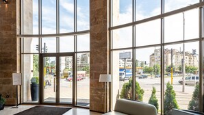 A spacious lobby with large windows, a curved sofa, and a round ottoman. - Hotel Vega Sofia (Sofia)