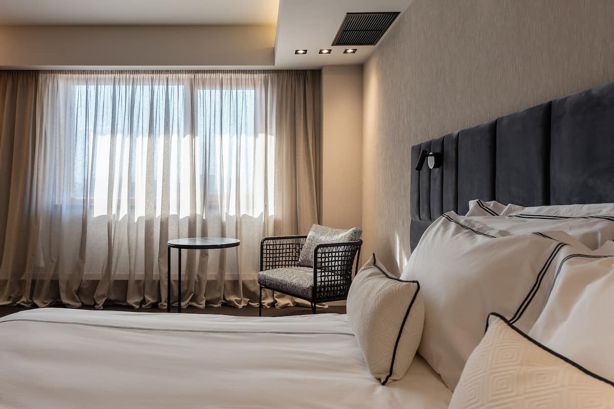 A hotel room with a bed, a chair, a small table, and a window with curtains.