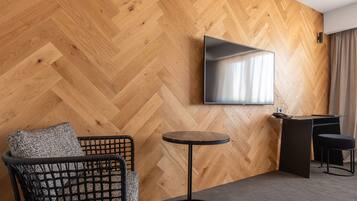 Una habitación con pared de madera en patrón de espiga, un televisor de pantalla plana sobre un escritorio, una mesa redonda y una silla negra con respaldo tejido.