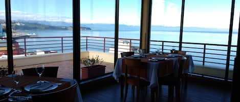 Un restaurante con vistas a un cuerpo de agua y montañas. Las mesas están dispuestas con copas de vino y platos.