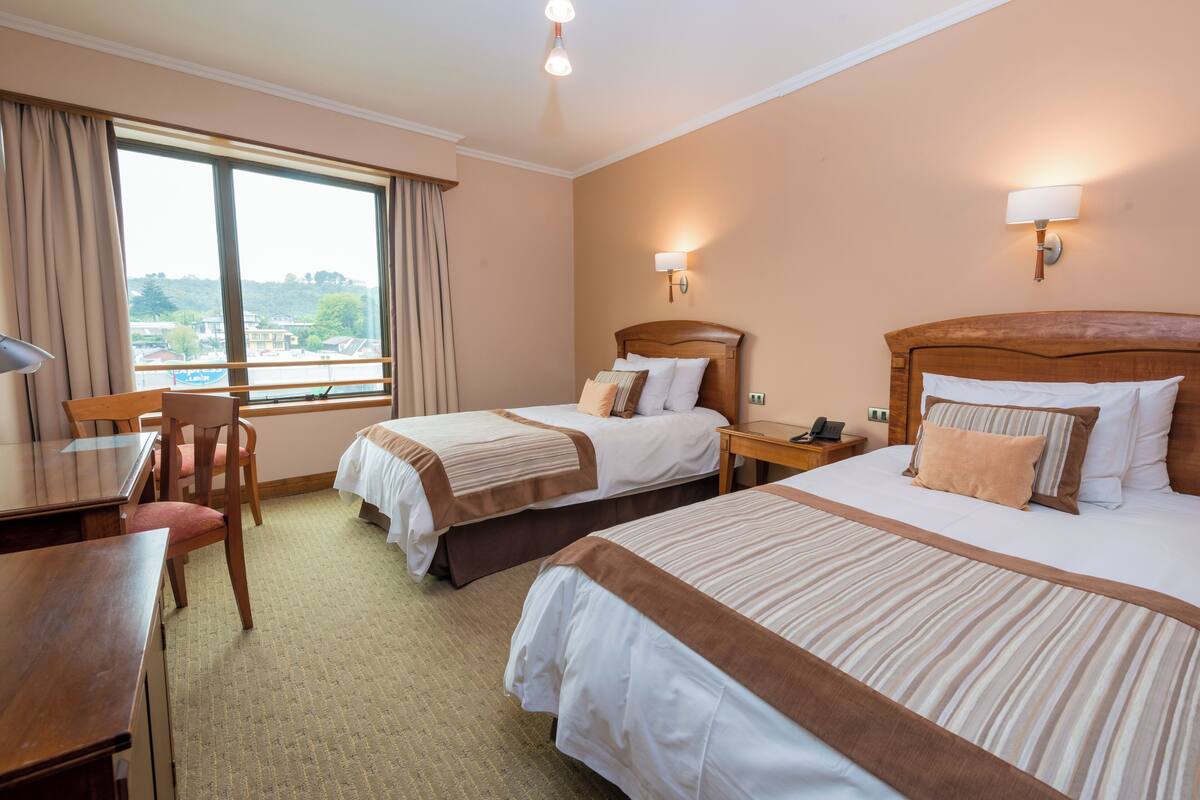 Habitación de hotel con dos camas, un escritorio y una silla. Incluye dos mesitas de noche, una lámpara y una ventana con cortinas.