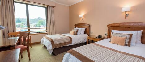 Quarto de hotel com duas camas, uma escrivaninha e uma cadeira. Há duas mesas de cabeceira, um abajur e uma janela com cortinas.