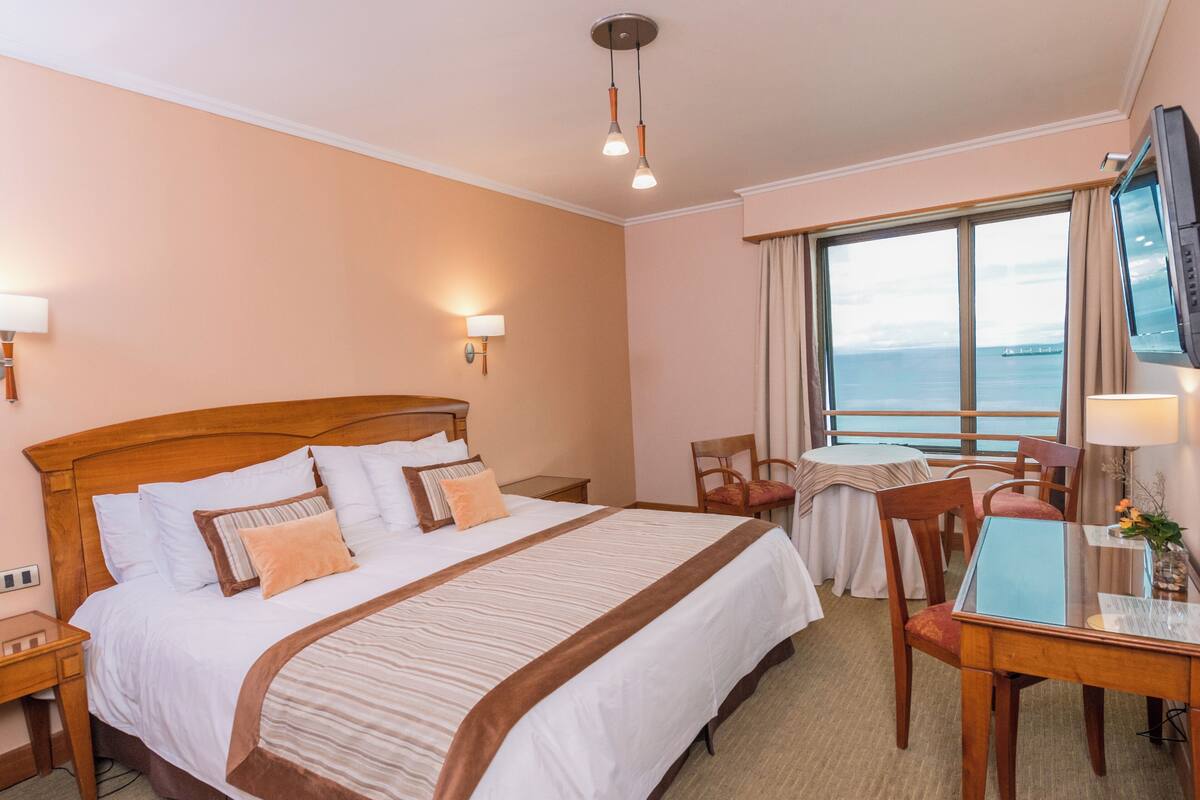 Habitación de hotel con una cama grande, un escritorio con sillas, un televisor y una ventana con vista al mar.