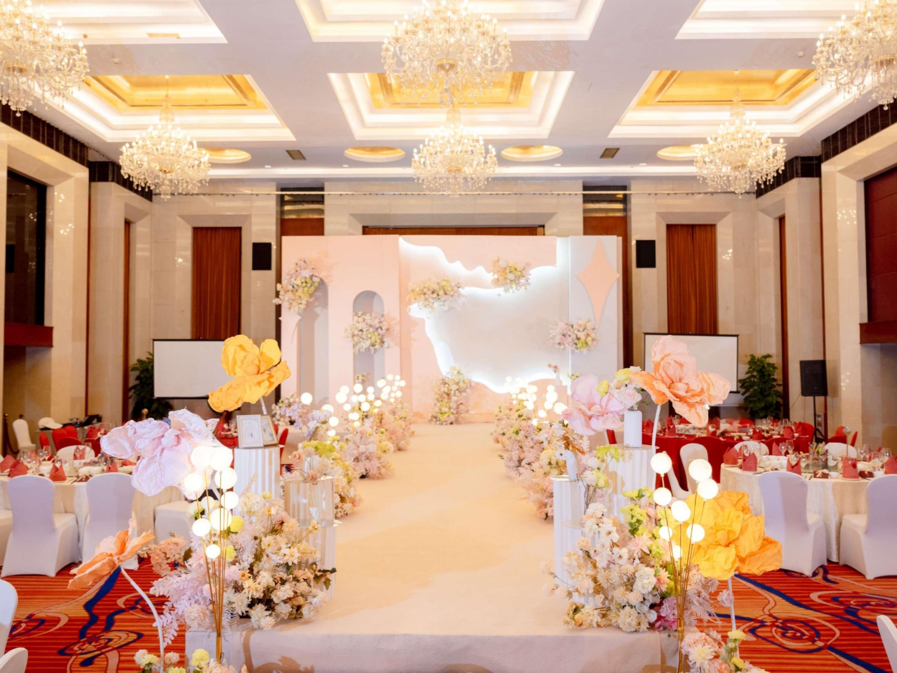 A banquet hall with a central aisle, floral decorations, chandeliers, and white chairs.