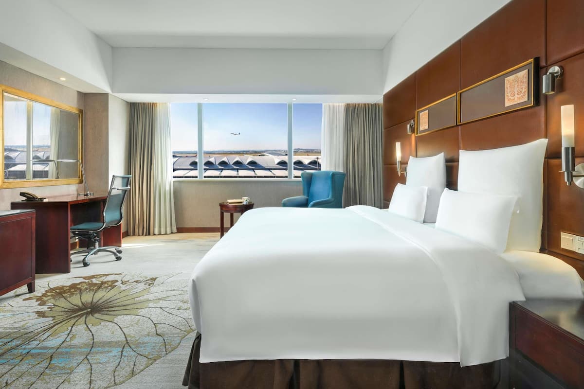 A hotel room with a large bed, a desk, a chair, and a view of an airport.