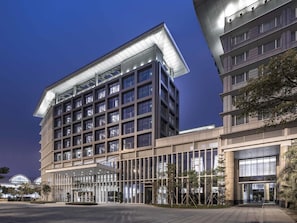 Un edificio de varios pisos con un diseño arquitectónico moderno, que destaca por su amplia fachada de vidrio y un área de entrada cubierta.