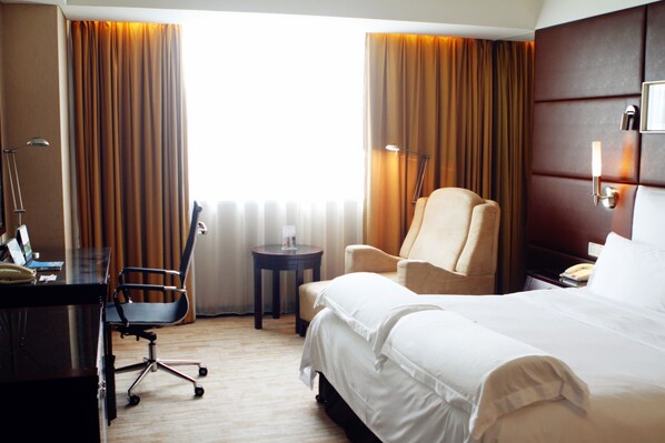 A hotel room with a bed, a desk, a chair, a lamp, and a view of the outdoors through curtains.