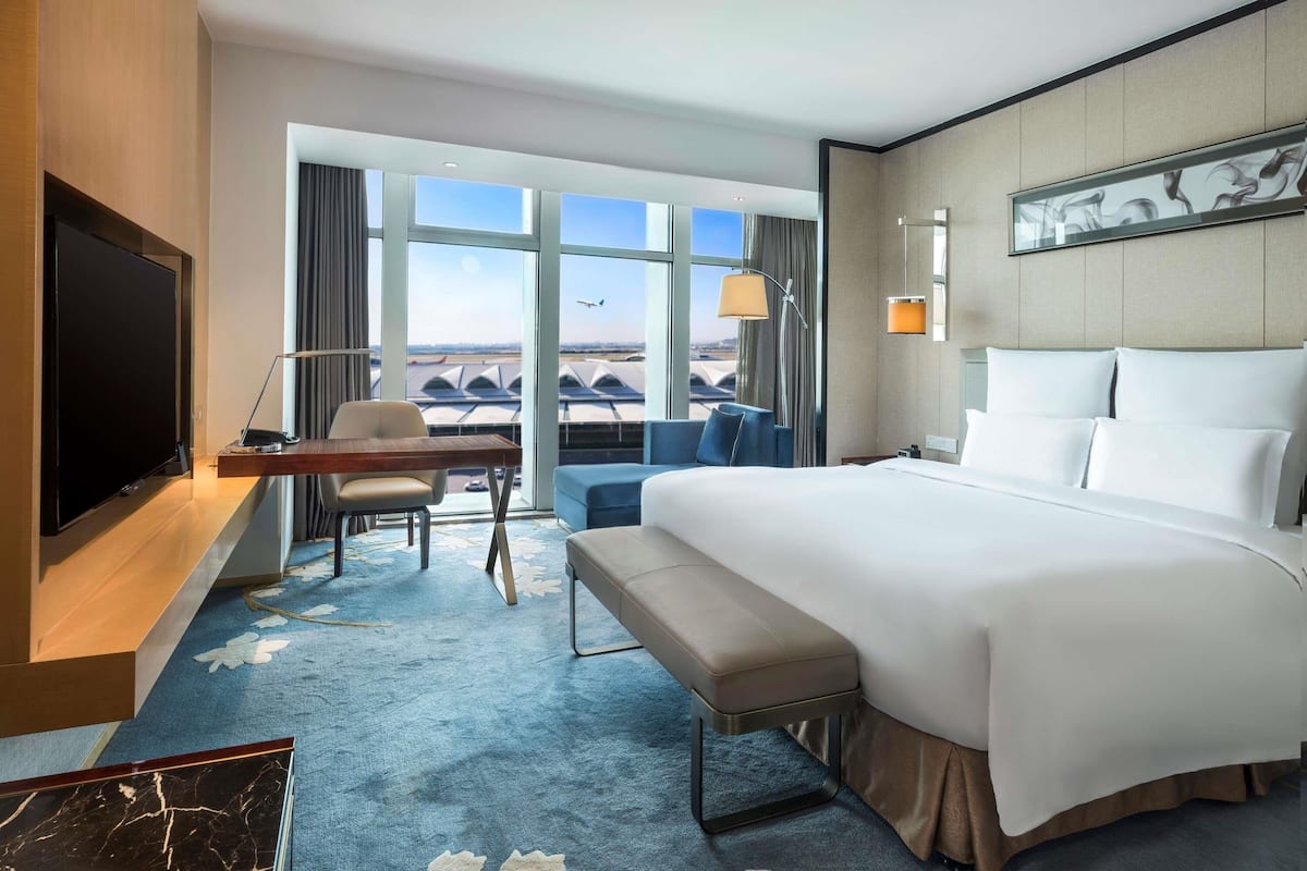 A hotel room with a large bed, a desk with a laptop, a chair, a bench, and a TV.