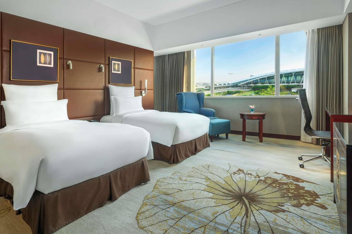 A hotel room with two beds, a chair, a small table, and a view of a modern building.