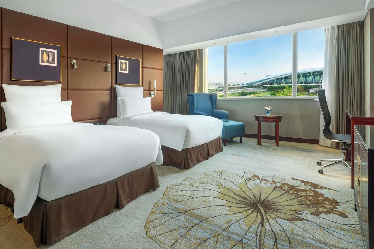 A hotel room with two beds, a desk, a chair, and a view of a bridge.