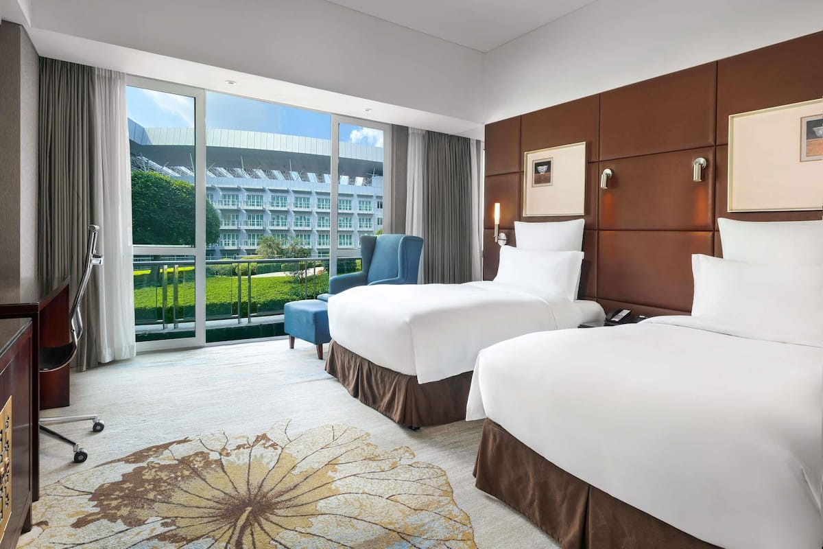 A hotel room with two beds, a desk, a chair, and a view of a modern building.