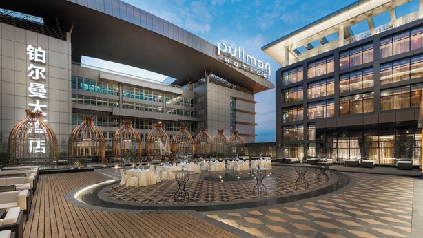 Un hotel de diseño moderno, con un amplio espacio exterior con mesas y sillas, una fuente central y un edificio grande con señalización.