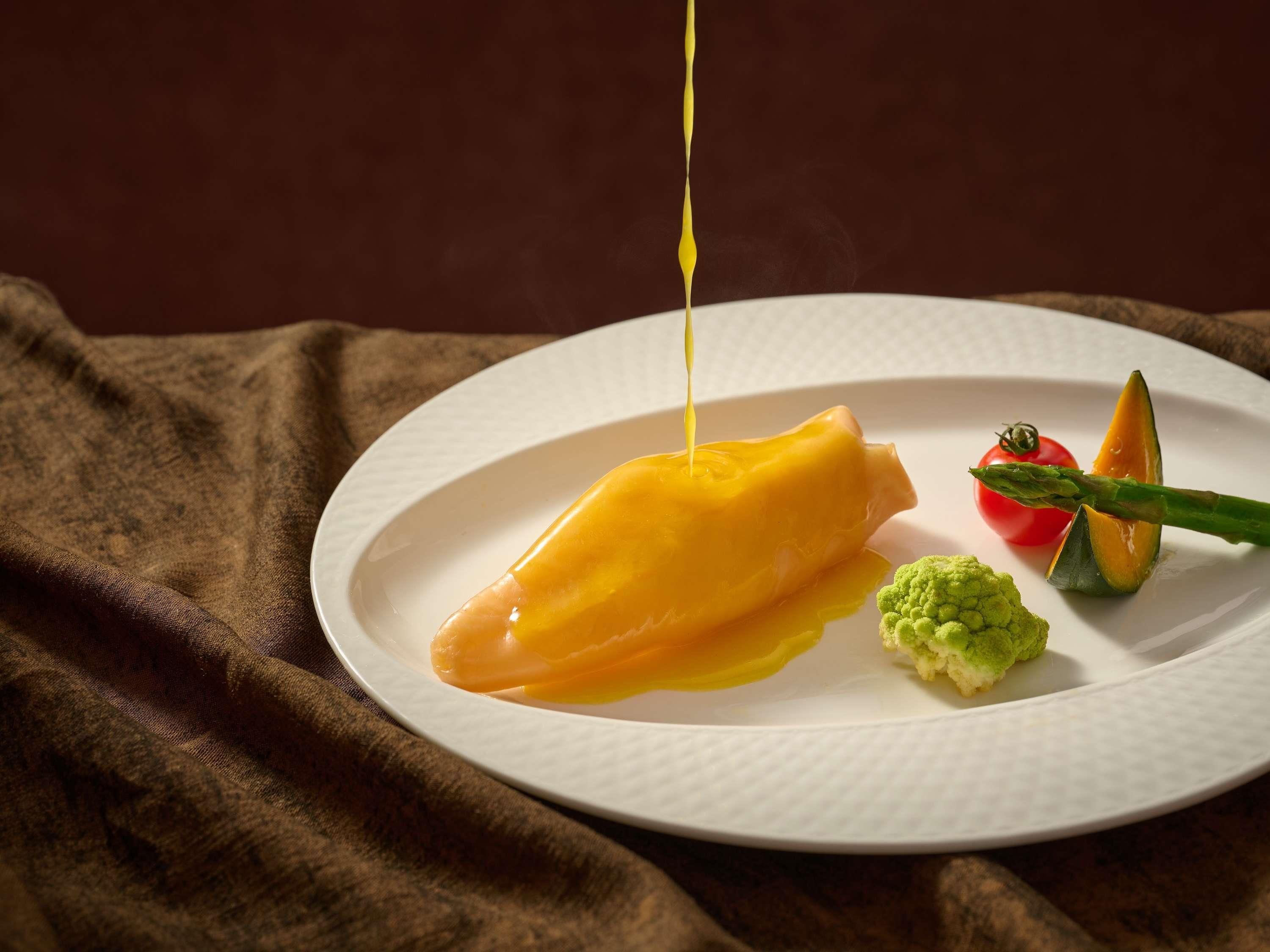 Un plato blanco con un postre amarillo, un tomate rojo y verduras verdes sobre un paño marrón.