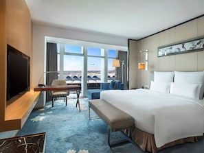 A hotel room with a large bed, a desk with a laptop, a chair, and a view of an airport.