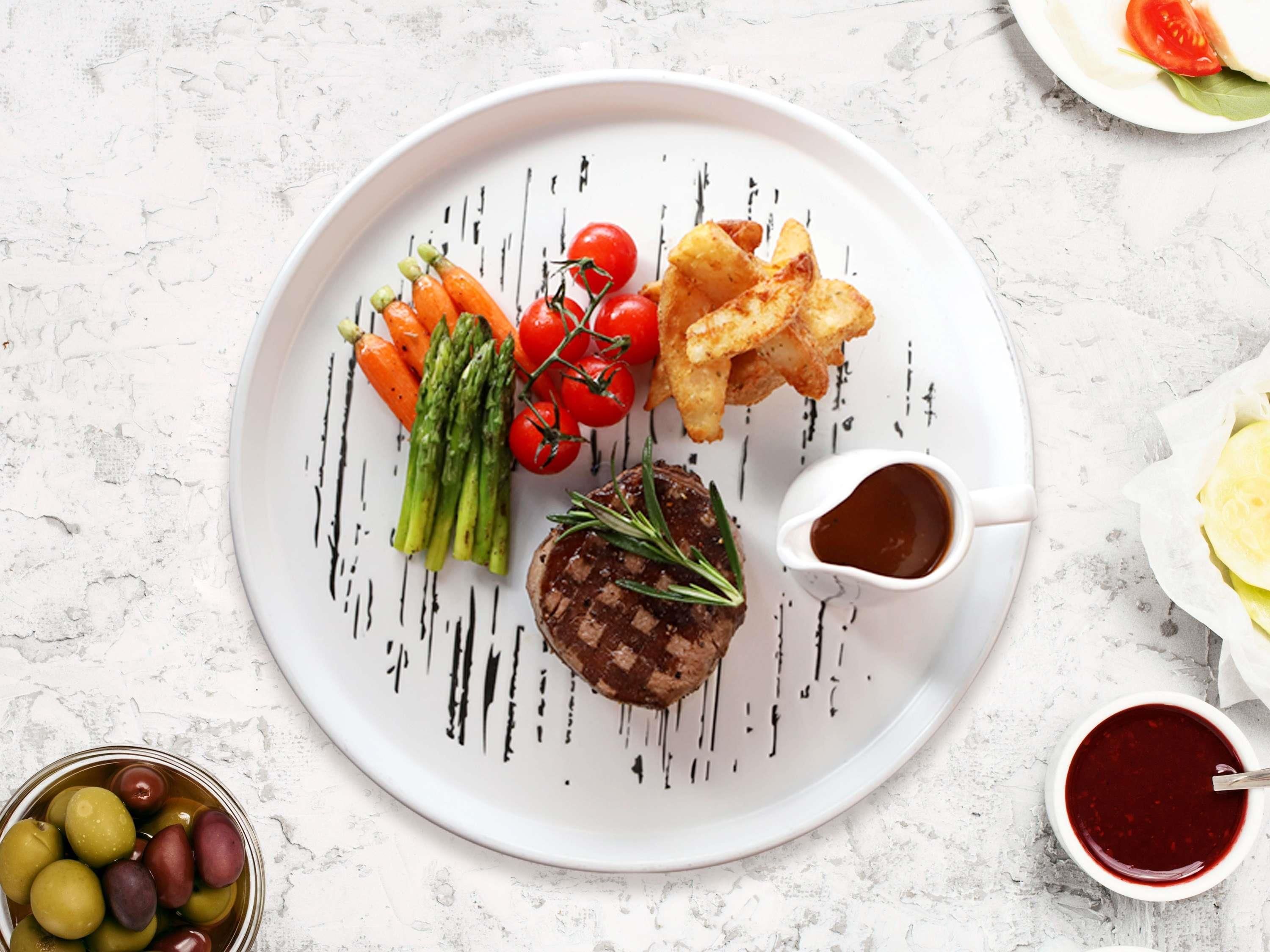 A plate with grilled steak, asparagus, cherry tomatoes, fried potatoes, and a small bowl of sauce.