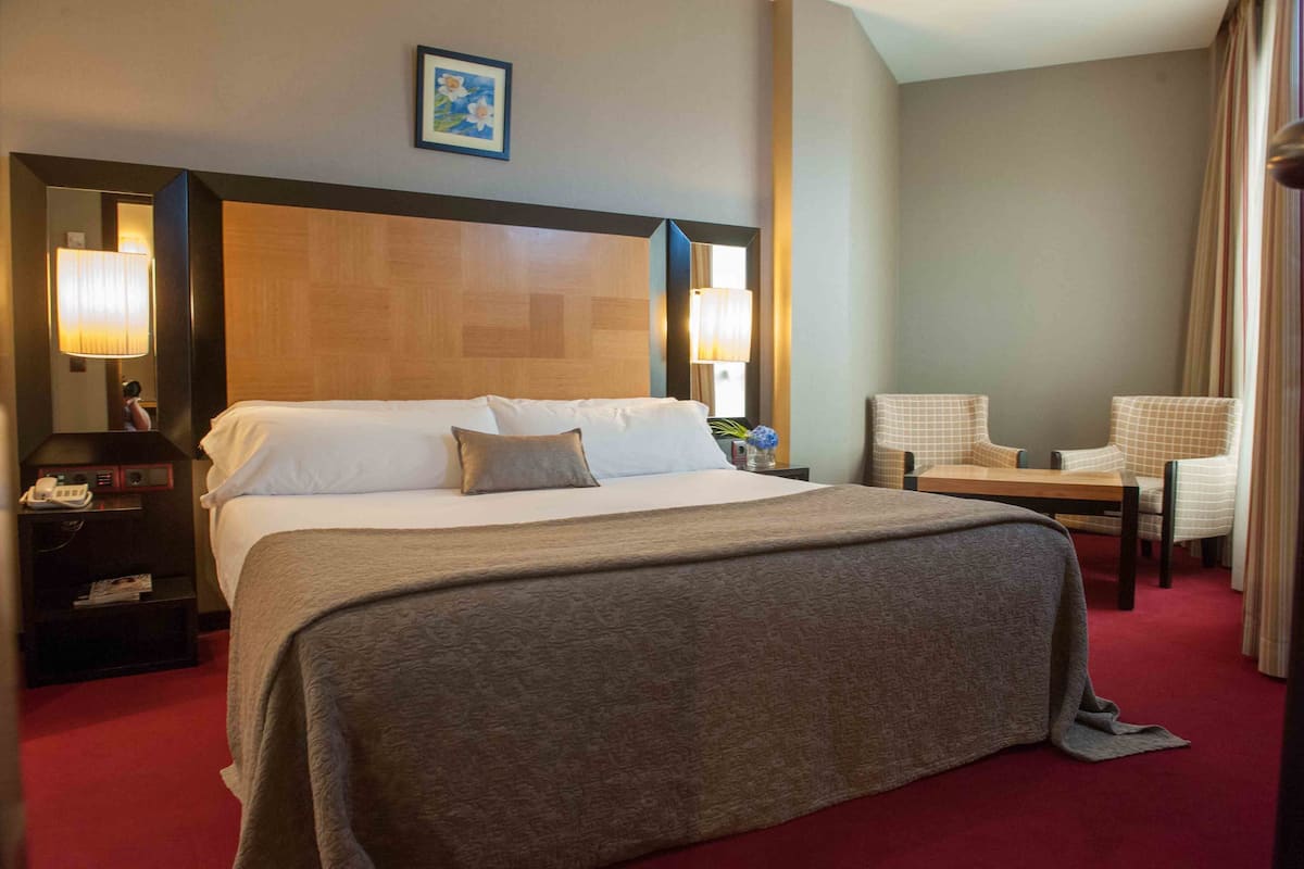 Una habitación de hotel con una cama grande, dos mesitas de noche con lámparas, una silla y una pequeña mesa.