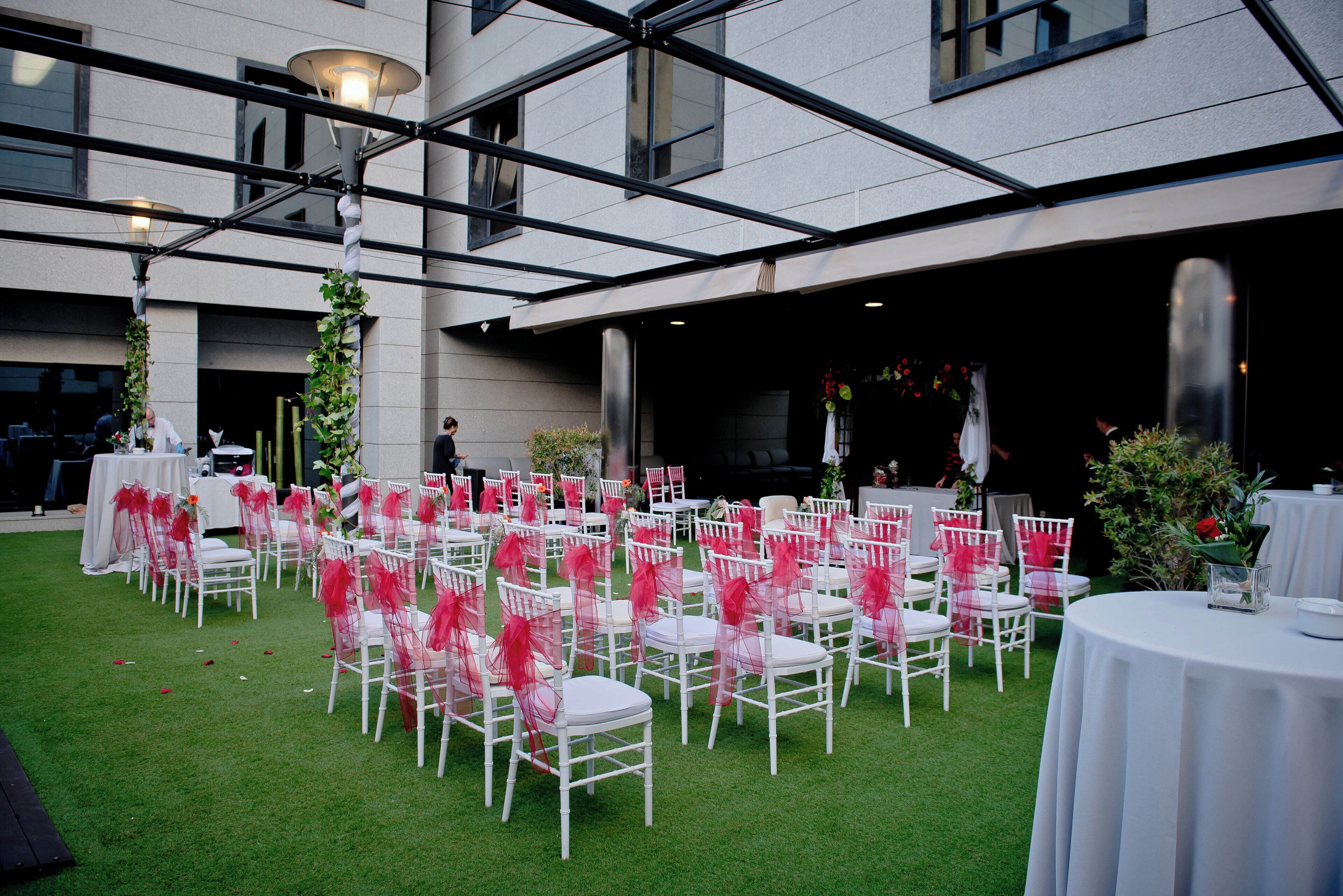 Un espacio para eventos al aire libre con sillas y mesas blancas, dispuestas en filas, y una zona cubierta con techo de cristal.