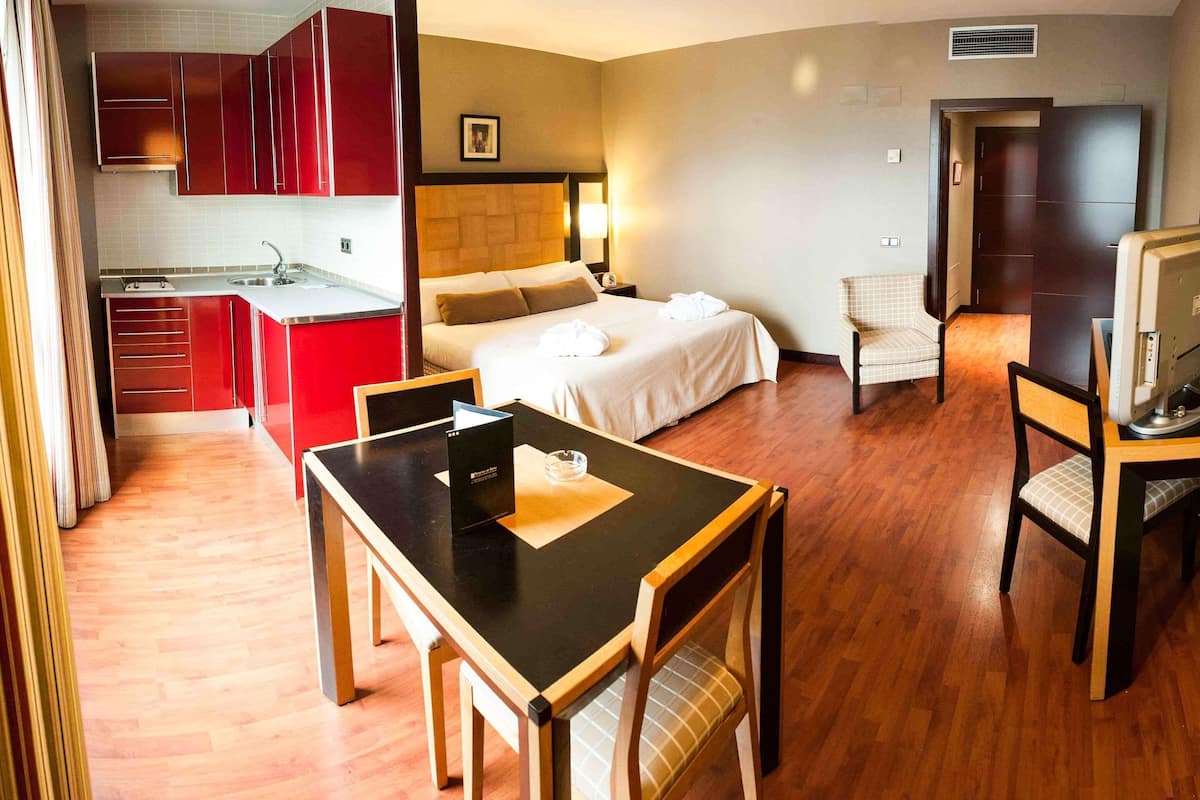 Habitación de hotel con cama, escritorio, silla, televisor, kitchenette y zona de estar separada.