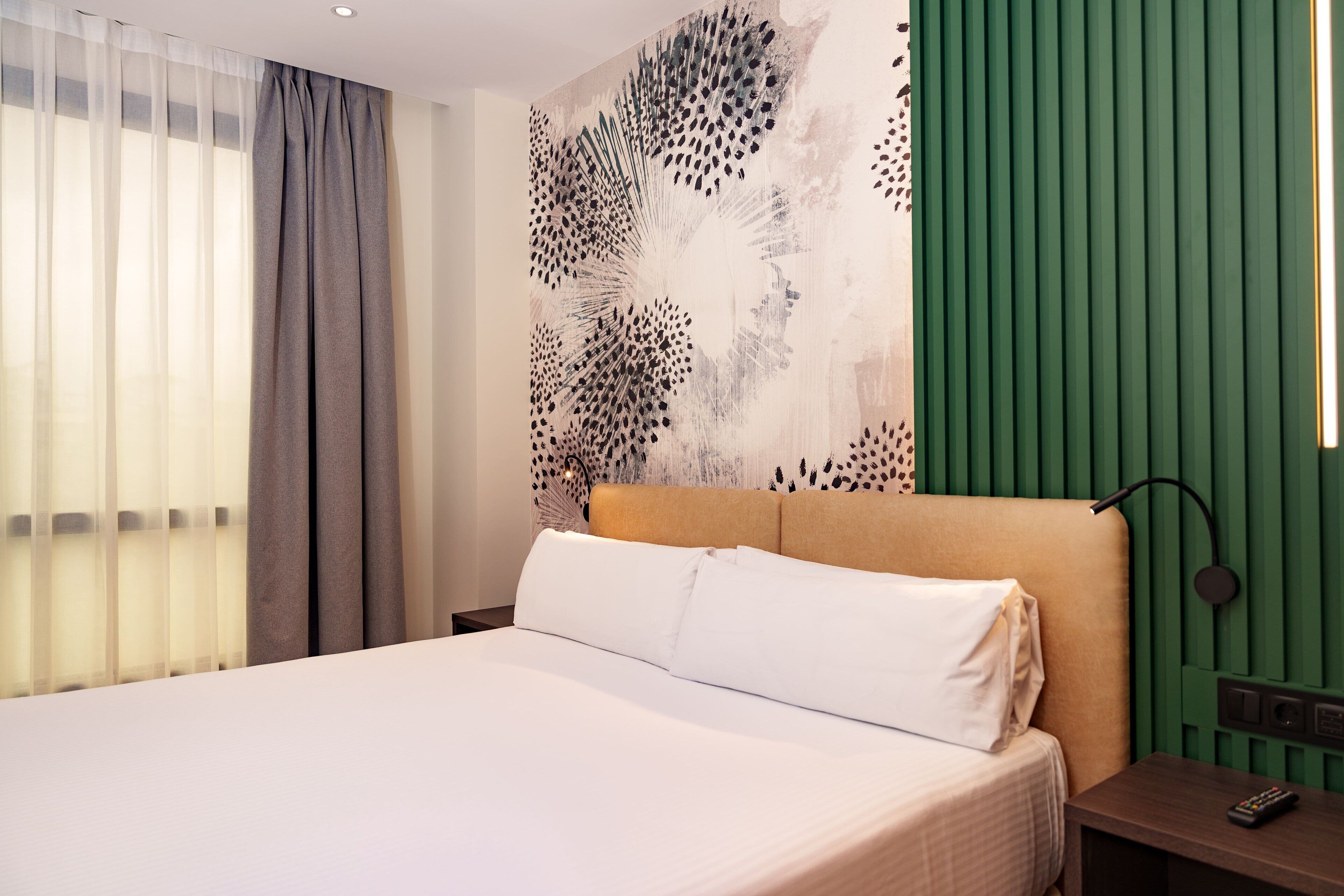 Une chambre d’hôtel avec un lit, une tête de lit, une table de chevet, une télécommande et un mur orné d’un panneau vert et d’un grand tableau abstrait.