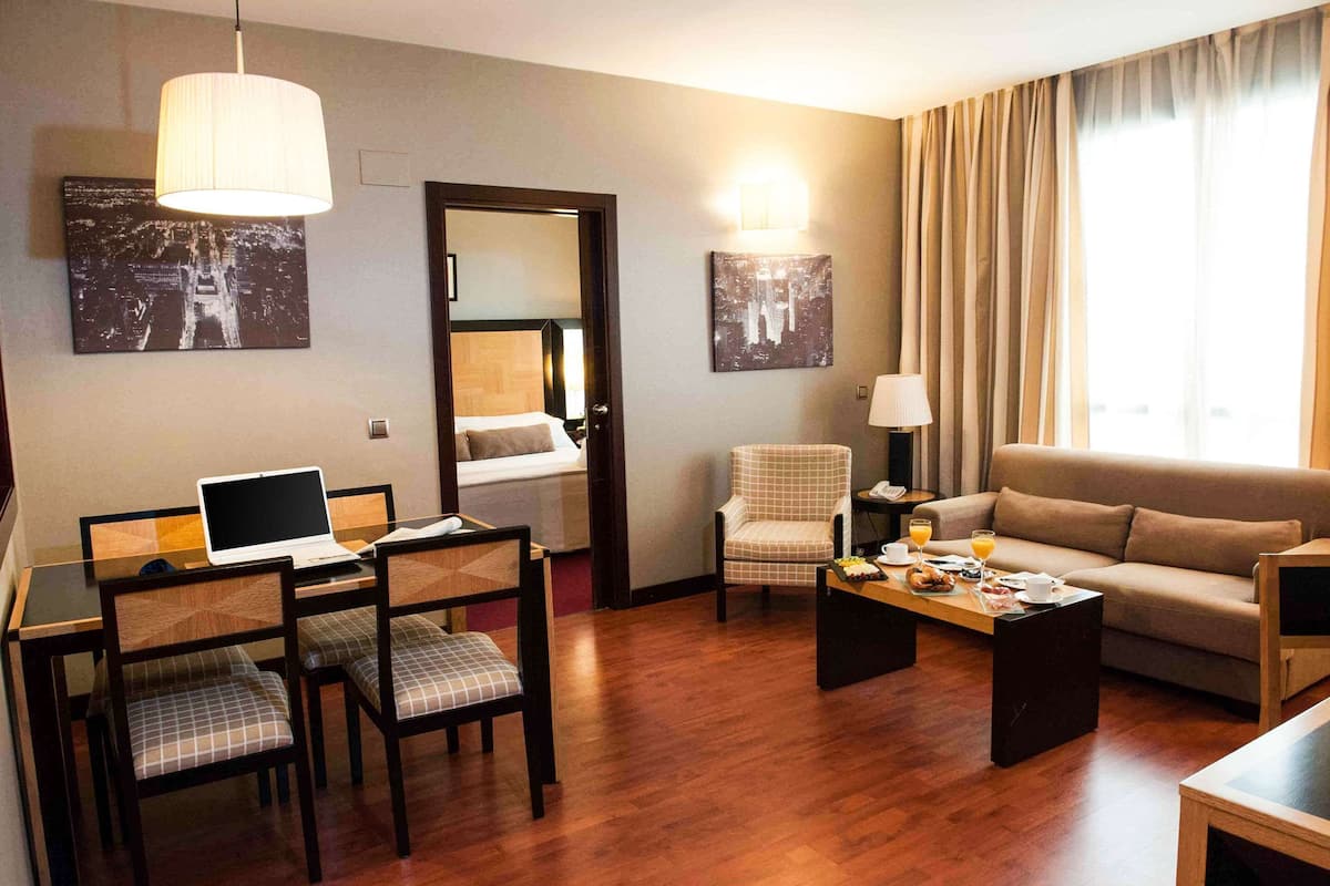 Habitación de hotel con mesa de comedor, sillas, una laptop, sofá, mesa de centro y dos lámparas de pared.