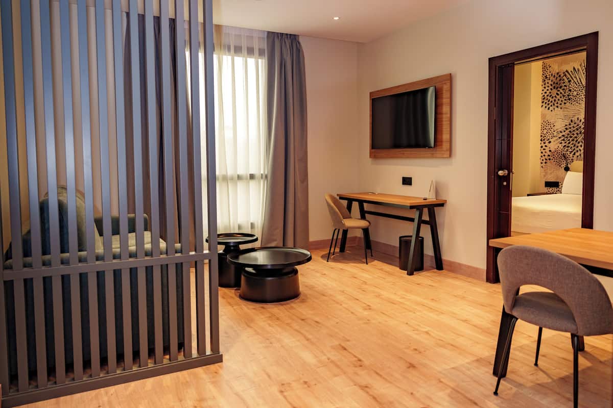Habitación de hotel con piso de madera, una cama, un escritorio con dos sillas, un televisor montado en la pared y una ventana con cortinas.