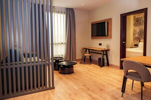 Habitación de hotel con piso de madera, una cama, un escritorio con dos sillas, un televisor montado en la pared y una ventana con cortinas.
