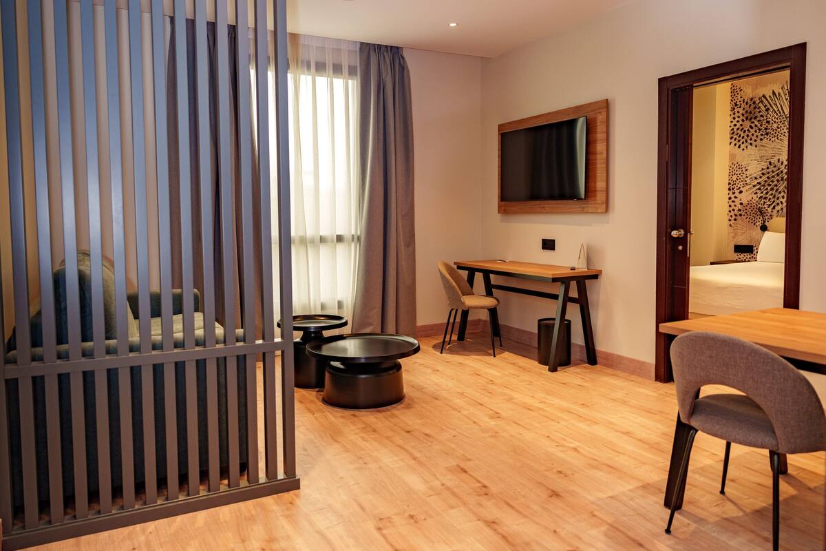 Una habitación de hotel moderna con suelo de madera, un sofá, una mesa de comedor con sillas, un televisor montado en la pared y un panel decorativo en la puerta.