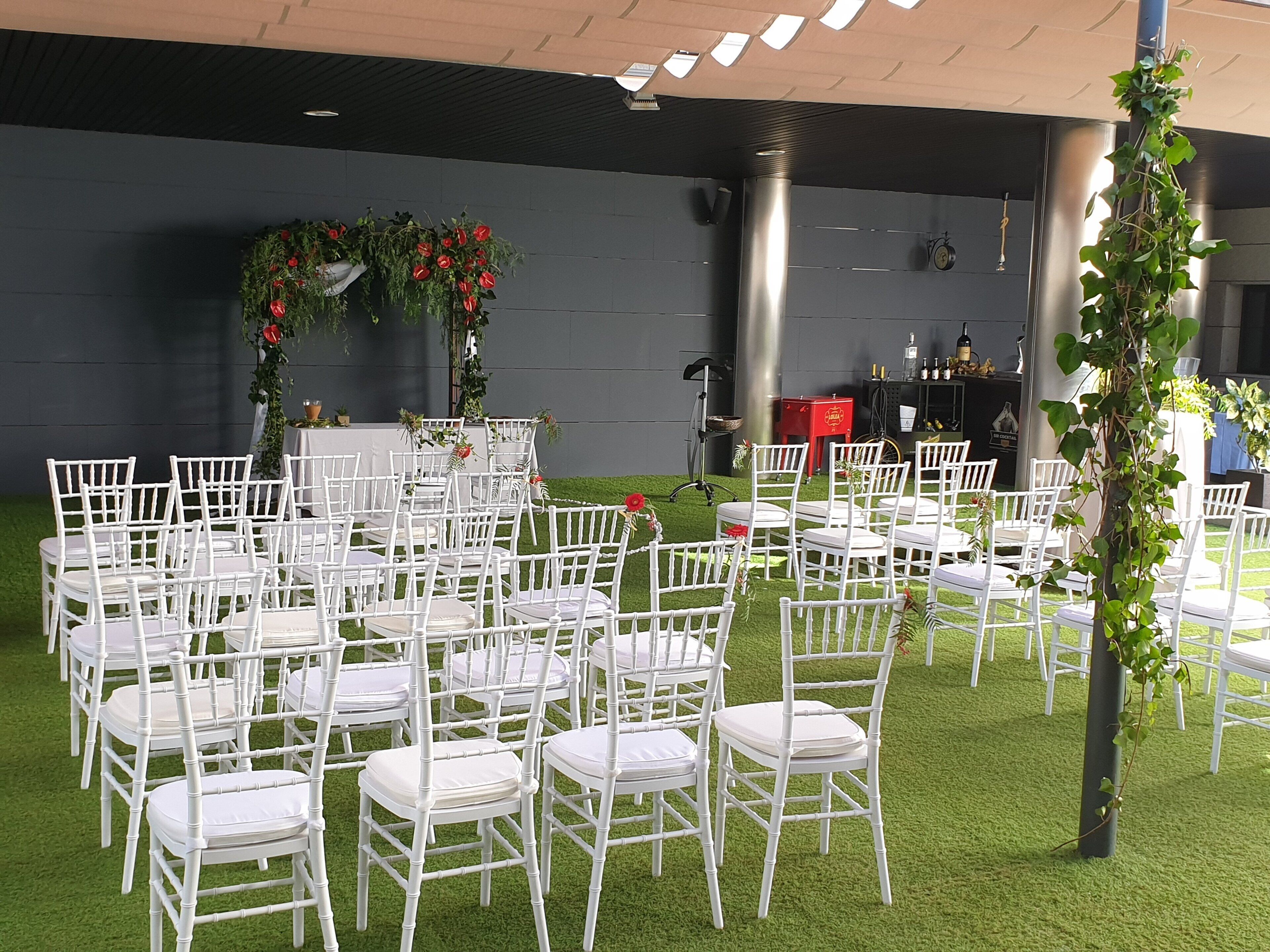 Un espacio para eventos al aire libre con sillas blancas dispuestas en filas, un escenario con decoraciones florales y una zona de bar con botellas.